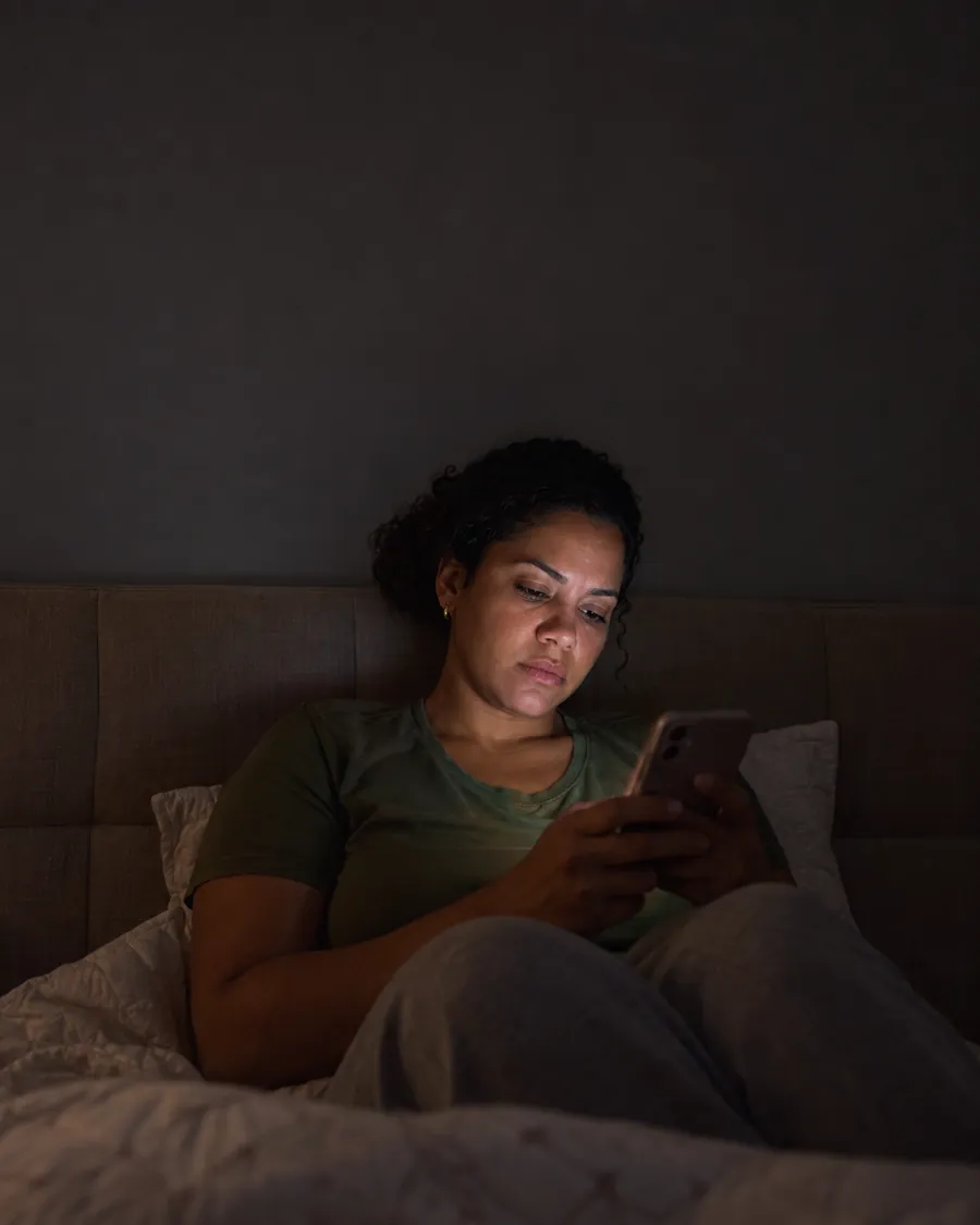 Imagem ilustrativa: mulher sentada na cama à noite, com o rosto iluminado pelo celular, olhando a tela com expressão cansada